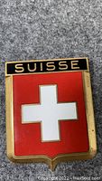 Close-up photo of the vintage Suisse enamel badge showing the red and white Swiss cross with 'SUISSE' text above, gold-tone metal border visible.