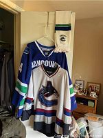 View showing two Vancouver Canucks NHL hockey jerseys on a hanger, one mostly blue and one white, and a Vancouver Canucks scarf hung over the top of the door.