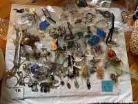 Overview of assorted jewelry items spread on a white cloth, with pieces in small plastic bags. Visible chains, earrings, pins, bracelets with mixed metals and colors.