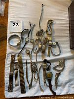 Photo shows all watches, bracelets and multiple metal watch bands laid out on fabric surface. Items vary in size and style, mostly gold or silver-tone metal with some leather bands.