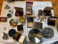 Top-down image of assorted vintage jewelry and accessory items laid out on cloth surface, showing various metal cuff links, belt buckles, jewelry boxes, compact mirrors, and pins.