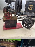 Photo showing the small steam engine model with brass body, red wheel, and attached red base alongside the fishing reel with dark metal and large open reel.