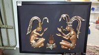 Full view of framed metal relief showing two figures and central fire on black background