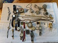 Full view of all watches, pocket watches, necklace chains and lockets laid out on a white cloth background showing variety of item types and condition.