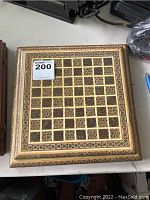 Top view of square wooden chess board with number 200 lot sticker placed on it. Shows the full board's inlaid geometric pattern.