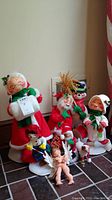 Group photo showing nine Annalee Christmas dolls and figures on tile floor against wall. Dolls include Mrs. Claus, elves, and children in holiday attire with holly decoration. Some dolls are on white stands. Dolls have bendable limbs and show some wear, needing cleaning.