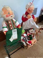 Photo showing Santa doll holding green sled and Mrs. Claus with white apron, along with small animal dolls on floor.