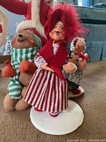 Close-up photo of the woman doll dressed in a red hood and striped skirt with a bear plush doll wearing a green and white striped scarf in the background.