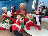 Seven Christmas-themed AnnaLee dolls arranged sitting and reclining on carpeted floor with blue backdrop, showing variety of Santa, elf, and female dolls with painted expressive faces and fabric clothing.