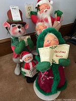 Five AnnaLee dolls including a green-hooded musical Caroler, three Santa figures in red and white, and a squirrel doll standing on the floor near a wall outlet