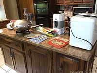 Kitchen island with various appliances: food processor, bread machine, iced tea maker, electric wok, grilling machine, and cutlery on countertop