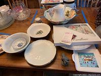 Display of the ceramic pitcher with floral bowl, several white plates, and small decorative items on a wooden table.