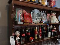 Wide view of multiple Coca-Cola collectibles on wooden shelves including bottles, figurines, truck model, polar bear cookie jar, paperweights, plate and framed art.