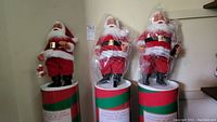 Three Coca-Cola Santa Claus figures standing on their original cylindrical boxes with red, green, and white Coca-Cola themed decorations, showing their full height and details.