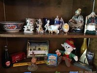 Shelf display with ceramic figurines, Goebel egg, Shirley Temple cup and pitcher, brass sailboat bookends, Ernie the Keebler Elf plush, Kentucky Derby glass, metal tray and other collectibles.