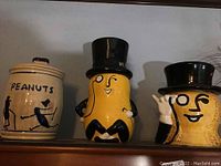 Three ceramic cookie jars including two Mr. Peanut shaped jars with black top hats and one tall jar labeled 'PEANUTS' with black silhouette figures.