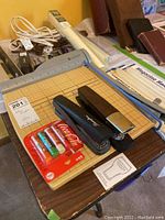 Photo showing assortment of office supplies including staplers, hole puncher, paper cutter, cords, and craft supplies on a table.