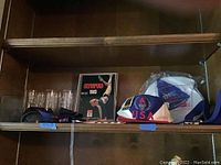 Shelf holding several Atlanta Olympics items including six drinking glasses, spiral bound 1983 Olympiad calendar and several Olympic hats including a packaged woman’s hat