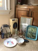 Photo showing a variety of Statue of Liberty themed items arranged on the floor near cabinet: decorative plates, glass, figurines, framed art, light in box, metal tray.