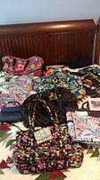 Wide image showing multiple Vera Bradley bags and accessories arranged on bed, including floral patterned fabrics and several bags.