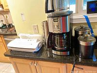 Cuisinart programmable 14-cup coffee maker with glass carafe and two stainless steel coffee mugs next to two canisters on countertop.