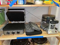 Front view of Cuisinart griddler with control knobs and open grilling surface, waffle and sandwich plates stacked beside it, and Waring Pro Belgian waffle maker on shelf.