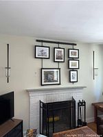 Wide view of black metal framed art system holding six framed artworks above a white fireplace, flanked by two metal wall sconces with glass holders.