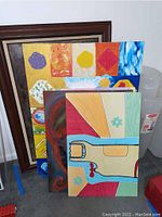 Four art pieces leaned against a wall including framed and unframed canvases and poster boards with colorful abstract and stylized designs.
