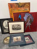 Photo showing a group of nine framed artworks and paintings leaning against the wall. Includes portraits, battle scenes, abstract and colorful paintings.