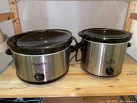 Two stainless steel Crock-Pot slow cookers side by side with black plastic trim and glass lids on wooden shelving.