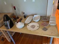 Table displaying electric wine bottle cooler, white ceramic teapot, Wedgwood Mrs. Tiggy-Winkle plate, assorted rectangular and divided ceramic serving dishes, some glass straws, and bottle-opening accessories.