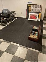 Overall view of the workout area showing the black interlocking rubber workout floor mat, duckpin bowling balls in wooden case, bowling shoes, weight bench, and framed pictures leaning on the wall.