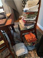 Stack of assorted linens folded on chair including napkins, place mats, bed linens in various colors and fabrics.