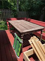 Outdoor wooden rectangular table with slatted top and turned legs, showing faded and weathered wood surface, placed on red deck corner bench seating.