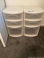 Two white plastic storage drawer units side by side on carpeted floor against a wall, each with three semi-transparent drawers.