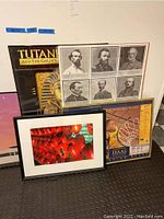 Collection of posters leaning against a wall including the framed lantern photo, TutanKhamun poster, generals poster without frame, basketball poster, and a partially visible poster.