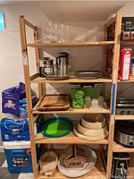 Photo of a wooden shelving unit holding multiple kitchen and serving items including stacked plates, wooden bowl sets, a fondue pot and stand, green decorative vase, glass containers, wooden platters, and baskets.