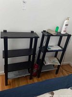 Two narrow black shelving units side by side against a white wall with various items on shelves.