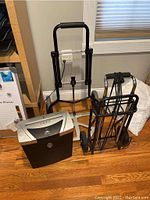 View of the shredder, luggage carrier, and folding hand truck together on wooden floor near window and cabinet.