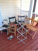 Four folding chairs (two natural wood, two black), two black step ladders, silver drying rack, and knee pads in package on a porch floor.
