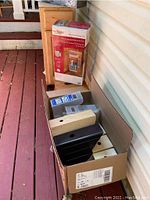 Overview showing wooden step stool, kitchen cabinet rough-in, boxed wood door storage unit, and various magazine holders (metal and wood) arranged on a red wooden floor against house siding.