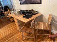 IKEA Norden drop leaf table with four wooden chairs, shown with some kitchenware on top. Table with solid natural wood finish and drop leaf sides. Wooden floor background.
