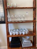 Wooden shelf with clear wine glasses on second shelf, white mugs on third shelf, and black and white square plates on bottom shelf.