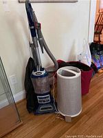 Photo of Shark Navigator vacuum and Swiffer Air Cleaner side by side on wooden floor, full view of vacuum showing hose and canister, and air cleaner cylindrical shape visible.