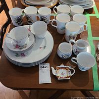 Photo of assorted china cups, mugs, saucers, plates, sugar bowl, creamer, and Ornate Orchid teaspoon holder arranged on table.