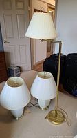 Three lamps: two table lamps with white glass bodies and brass bases, and one brass adjustable floor lamp with white pleated shade, seen in a room with beige carpeting.