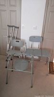 Photo shows two gray aluminum mobility aids with perforated seats and backrests, one with three seat panels and armrest, other a single wide seat, along with pair of aluminum crutches leaning against the wall.