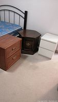 Three night tables grouped beside a bed, shows different styles and colors including dark brown octagonal, medium brown two-drawer, and small white two-drawer tables.