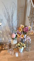 Tall ceramic vase with faux dried branches and pink rose, surrounded by multiple clear glass jars and vases. Artificial flower arrangements include pink and yellow calla lilies, roses, and small yellow roses.