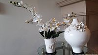 Two white ceramic vases on a glass table in front of a wall, one taller with bulbous shape and floral decorations, smaller one with faux orchid flowers.